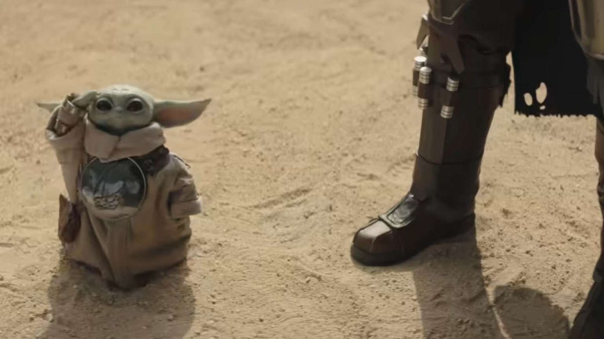 Baby Yoda (Grogu) in beige robe waves on a sandy desert with an armored figure's leg nearby.