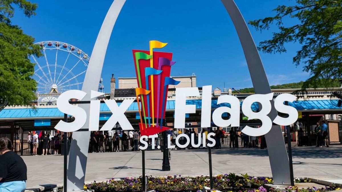 Six Flags St. Louis entrance sign with a colorful vertical sculpture and a Ferris wheel in the background on a sunny day.