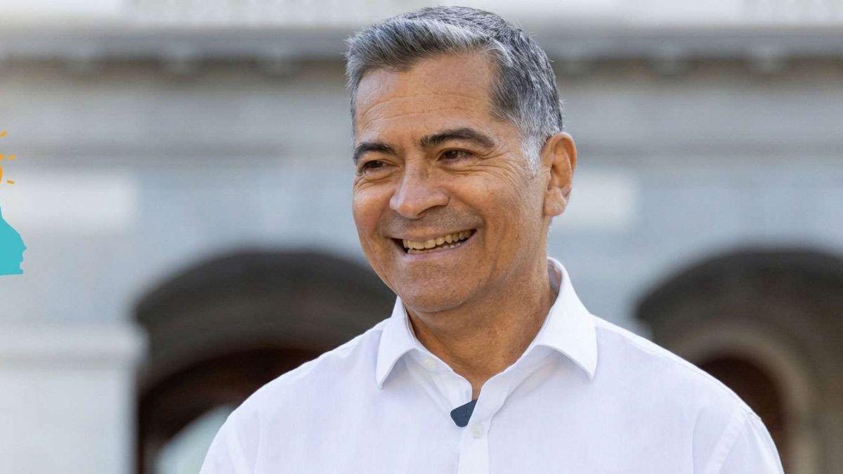 Smiling middle-aged man with short gray hair in a white dress shirt, outdoors against a light background.