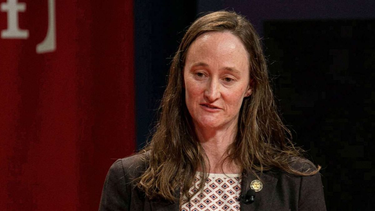Professional woman in a dark blazer giving a talk, wearing a patterned blouse and a circular pin on her lapel.