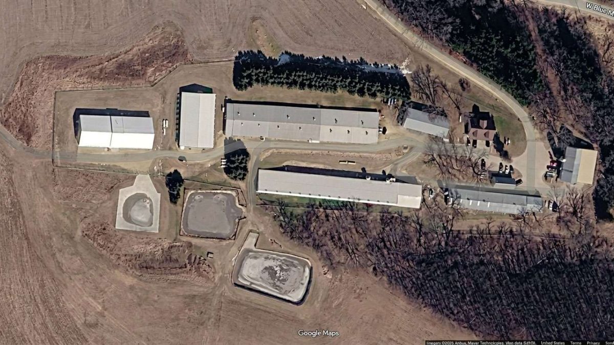 Aerial view of a rural industrial site with several long rectangular buildings, a parking area, and dirt roads among leafless trees.