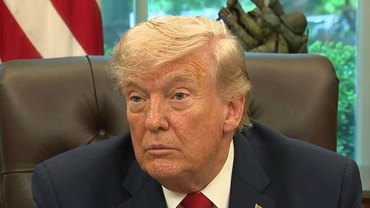 Man in a dark suit and red tie seated in a leather chair at a desk in an office, with an American flag in the background.