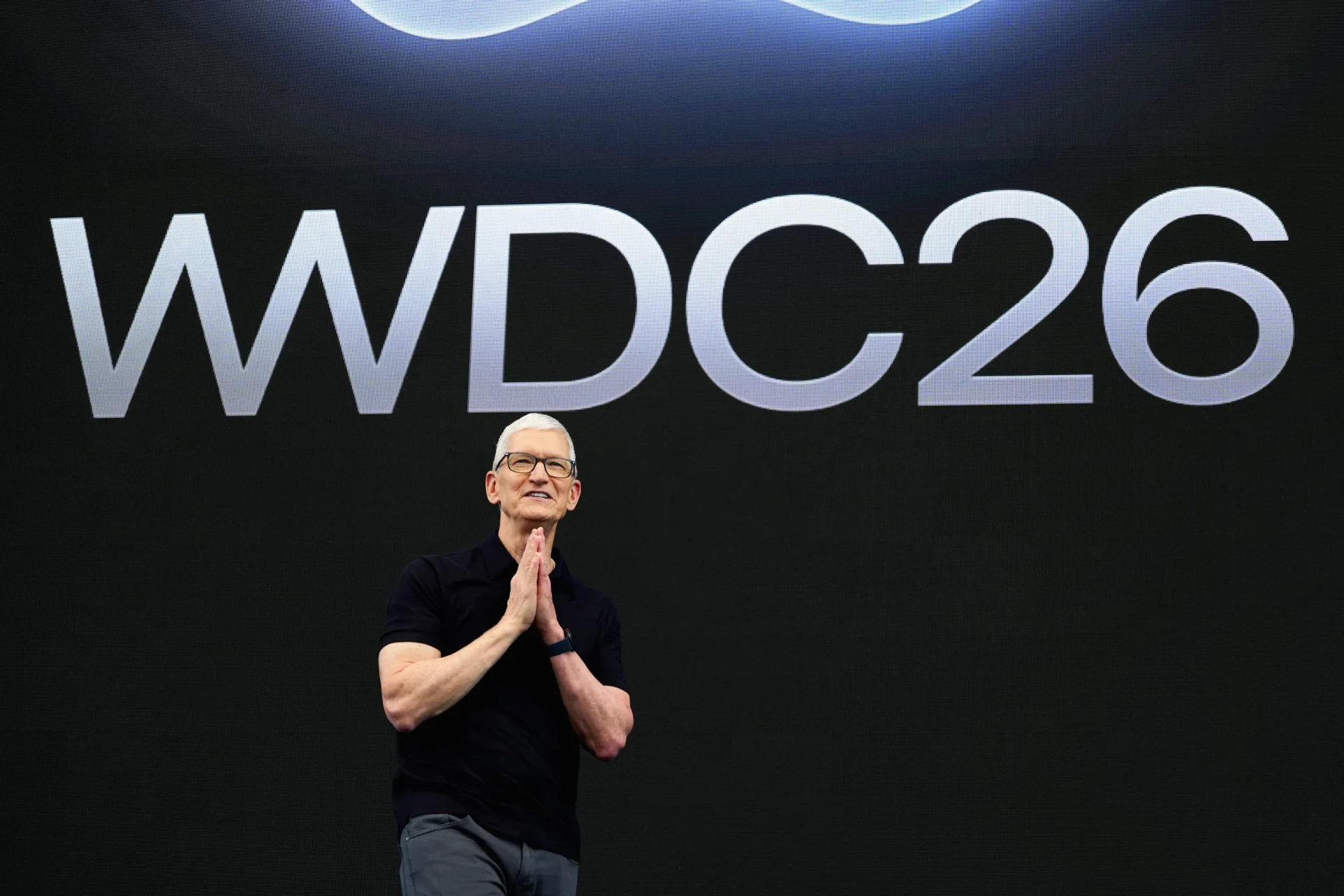Man in a dark shirt on stage with hands pressed together, large WWDC26 text on a black backdrop behind him.